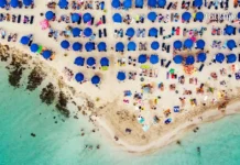 დაუვიწყარი ტური კვიპროსის სანაპიროზე – აია ნაპა გელოდება! Top-down aerial view of a crowded tropical beach with clear water and blue umbrellas.