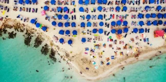 დაუვიწყარი ტური კვიპროსის სანაპიროზე – აია ნაპა გელოდება! Top-down aerial view of a crowded tropical beach with clear water and blue umbrellas.