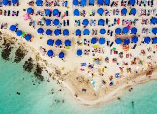 დაუვიწყარი ტური კვიპროსის სანაპიროზე – აია ნაპა გელოდება! Top-down aerial view of a crowded tropical beach with clear water and blue umbrellas.