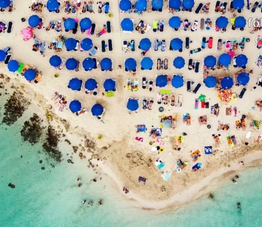 დაუვიწყარი ტური კვიპროსის სანაპიროზე – აია ნაპა გელოდება! Top-down aerial view of a crowded tropical beach with clear water and blue umbrellas.