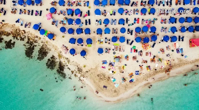 დაუვიწყარი ტური კვიპროსის სანაპიროზე – აია ნაპა გელოდება! Top-down aerial view of a crowded tropical beach with clear water and blue umbrellas.